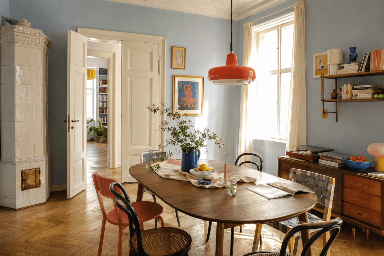 Sunlit dining room with pale blue walls, a red pendant light, colourful art, and mixed chairs